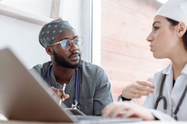 Two healthcare colleagues in scrubs and a white coat talking beside a laptop