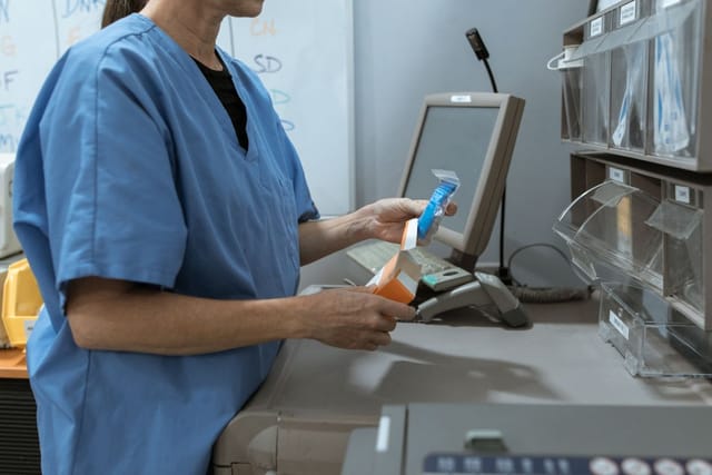 Healthcare worker in blue scrubs examining a medicine package beside dispensary shelves